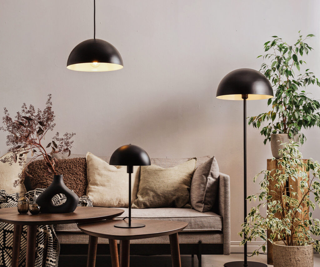 Taklampe, gulvlampe og bordlampe i stue. Sorte lamper, beige sofa og en stor plante til høyre for sofaen.