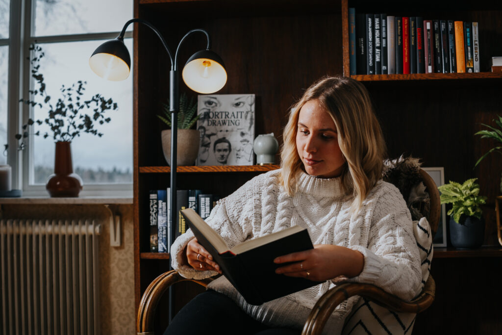 En kvinne sitter i en lenestol i stuen og leser i lyset fra en svart gulvlampe.